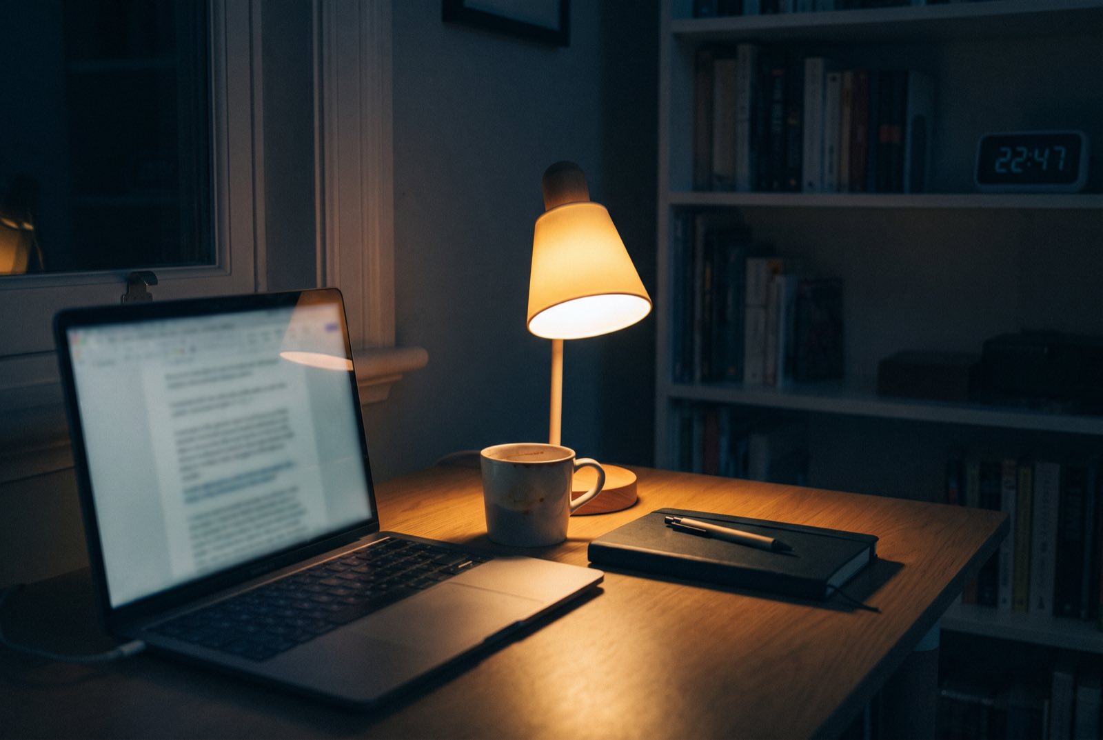 Escrivaninha à noite: laptop aberto com um documento pela metade, luminária acesa e o relógio marcando 22:47.